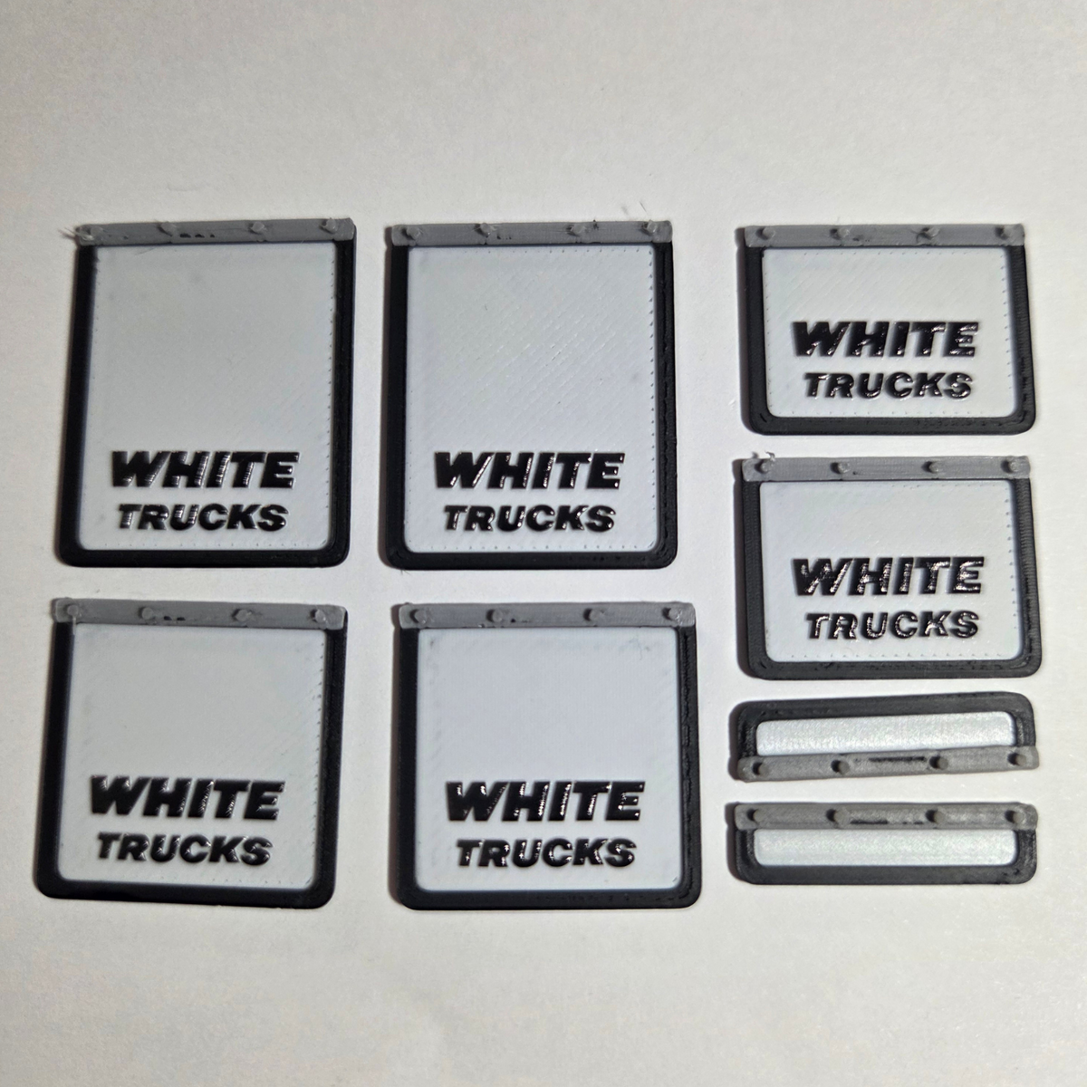 Six white rectangular scale model mudflaps with black trim and the words 'WHITE TRUCKS' printed on them, arranged on a white background with a black border and text reading 'SCALE MODEL MUDFLAPS White Trucks'.